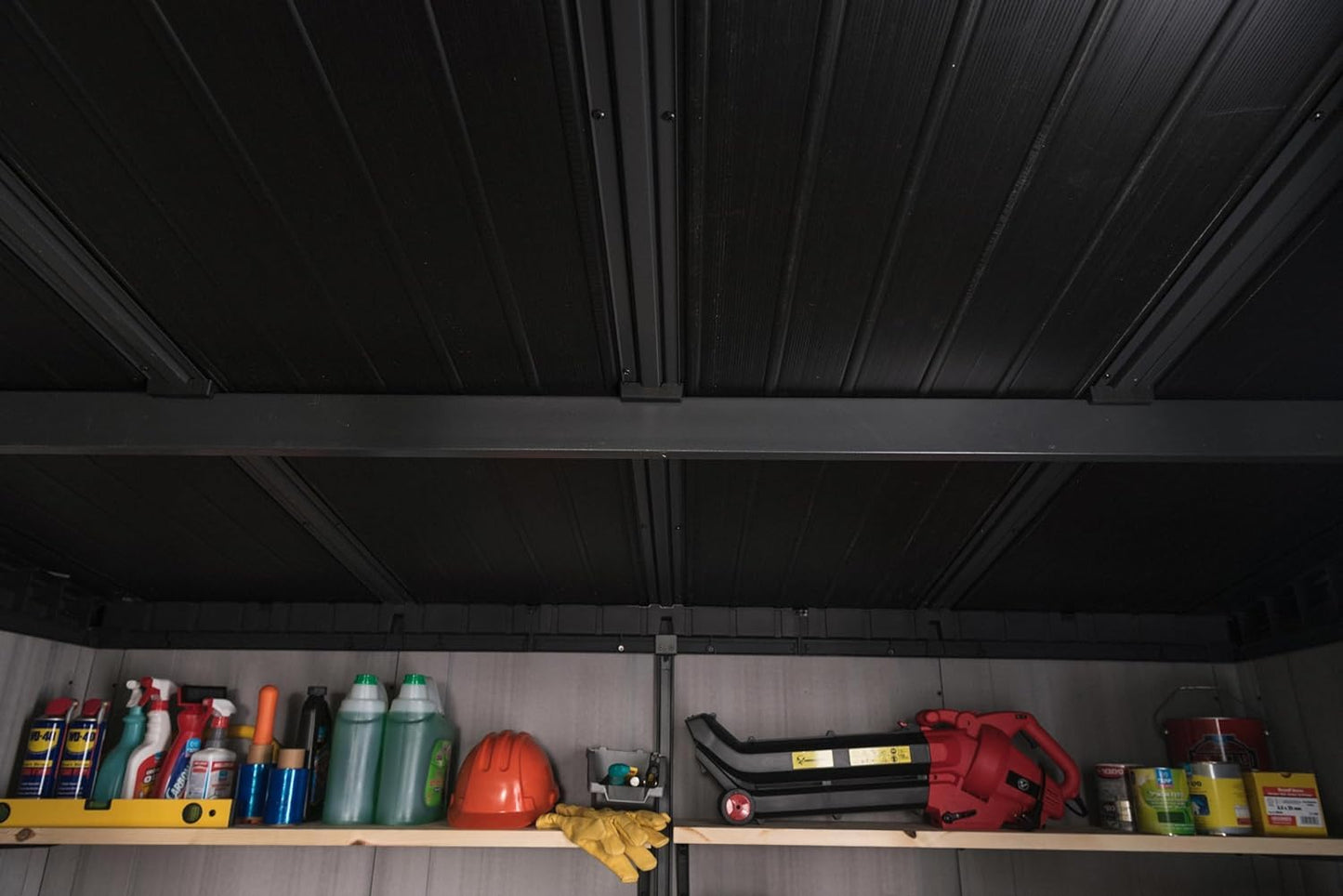 Interior view of shed ceiling with black support beams and a top shelf holding bottles, a helmet and tools.