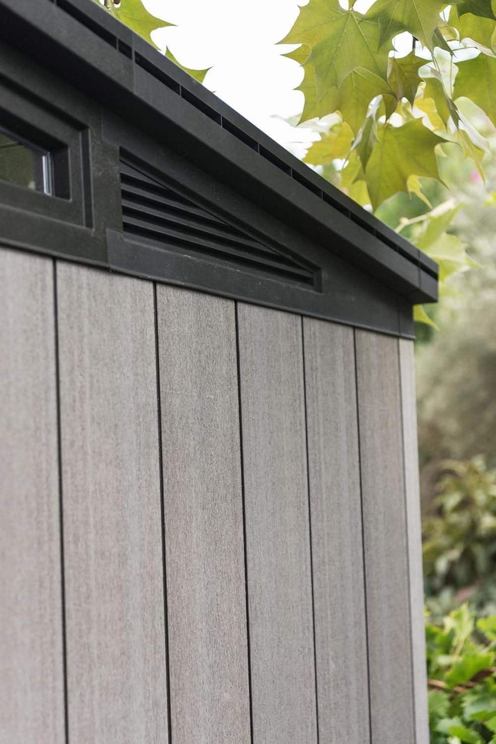 Close-up of roofline ventilation grille above grey wood‑effect panels with tree leaves visible
