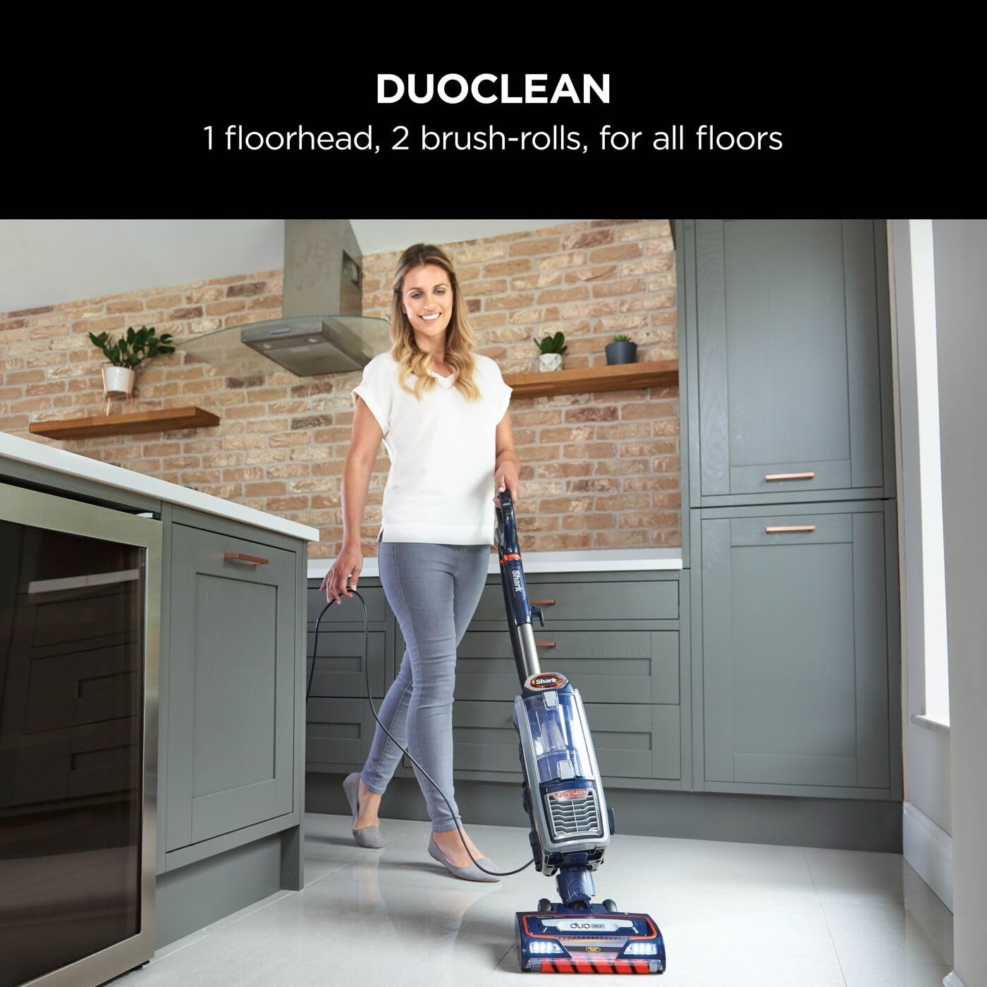 Person pushing a Shark upright vacuum across a tiled kitchen floor, demonstrating the DuoClean floorhead in use.