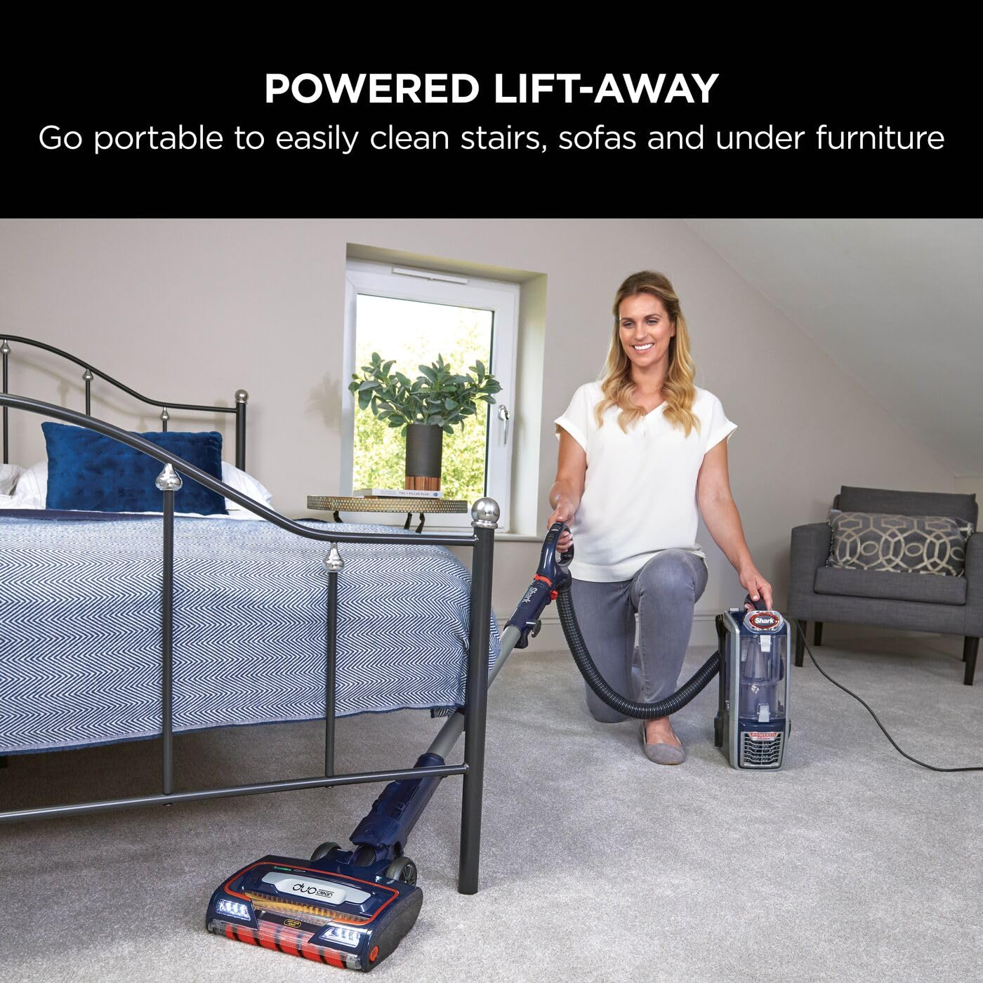 Woman using the Lift-Away portable canister and hose to vacuum a bedroom floor beside a bed.