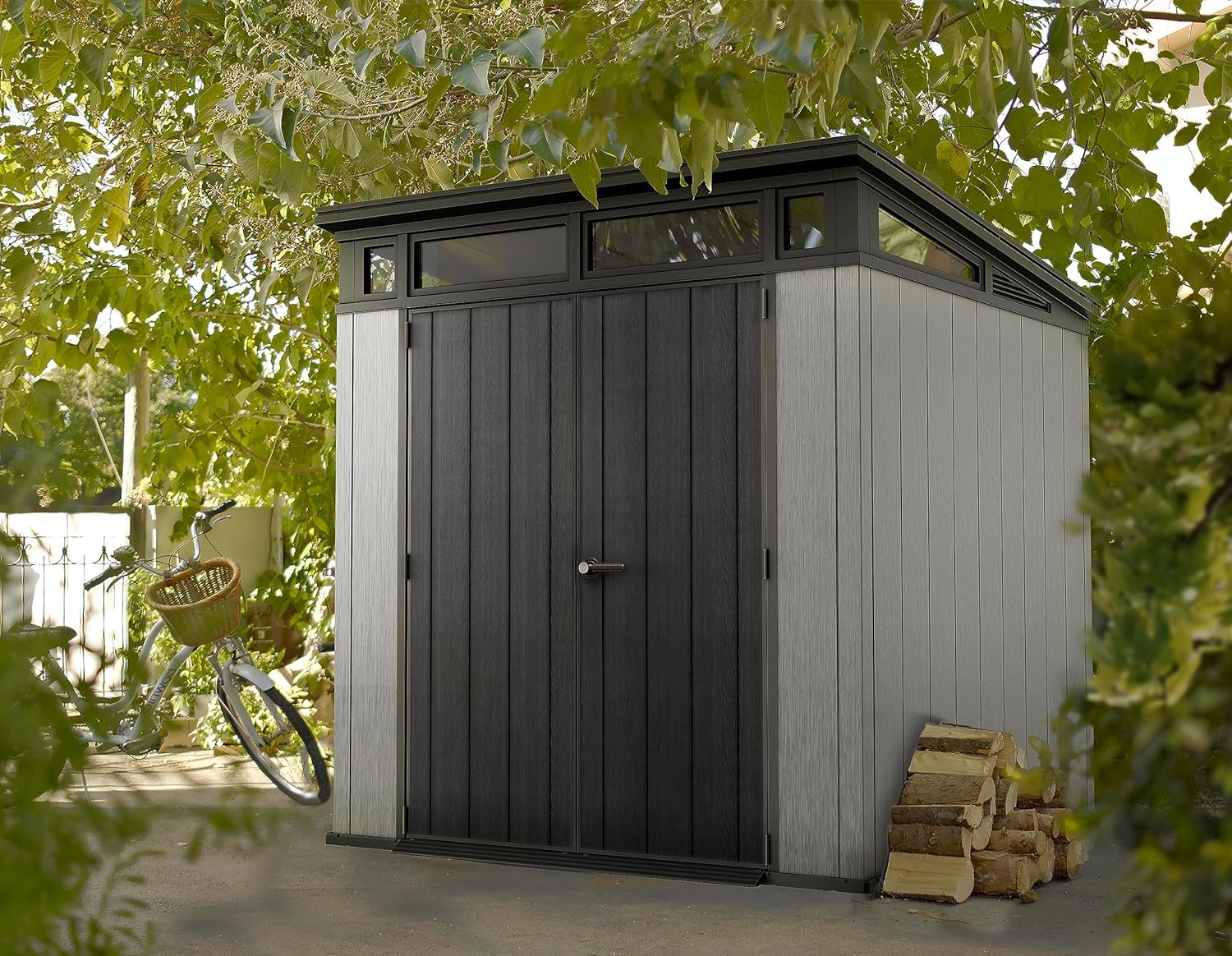 Front view of closed grey-and-black shed framed by trees, with a bicycle and stacked firewood beside it.
