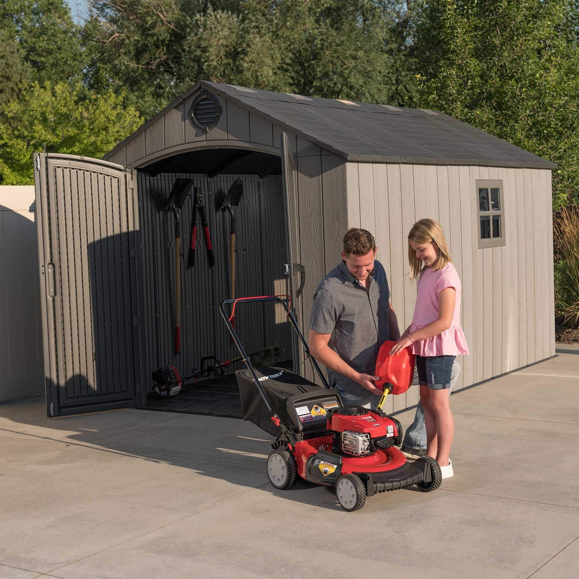 Open shed with double doors ajar revealing stored tools while a man and child handle a red lawn mower outside.