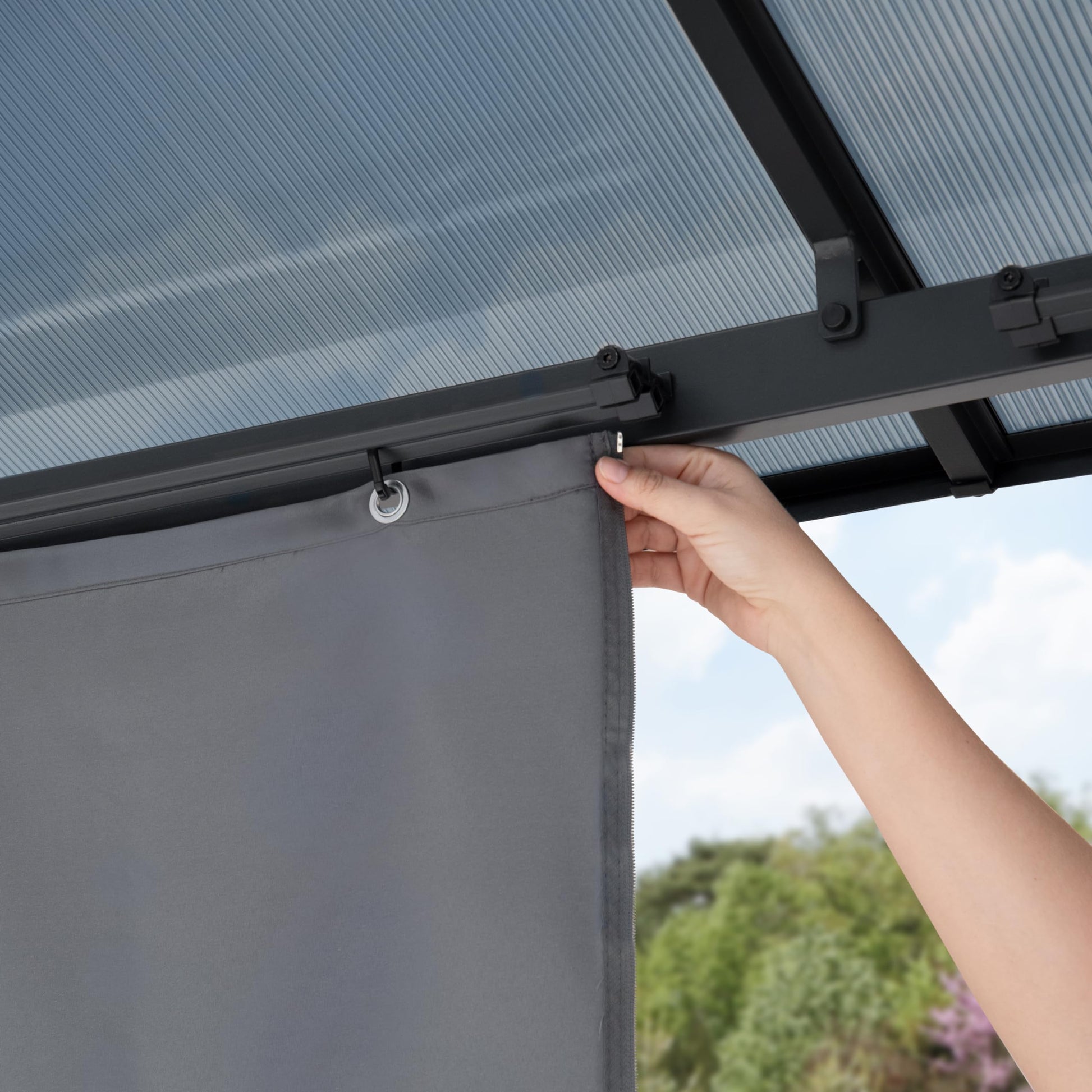 Detail of ribbed polycarbonate roof panels and a hand attaching a curtain hook to the metal frame.