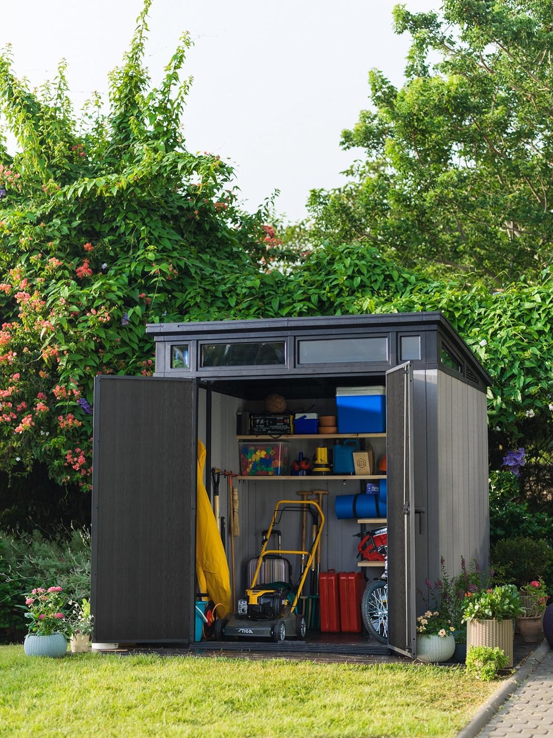 Wide exterior view of open garden shed filled with tools, a lawnmower and shelves, surrounded by plants.