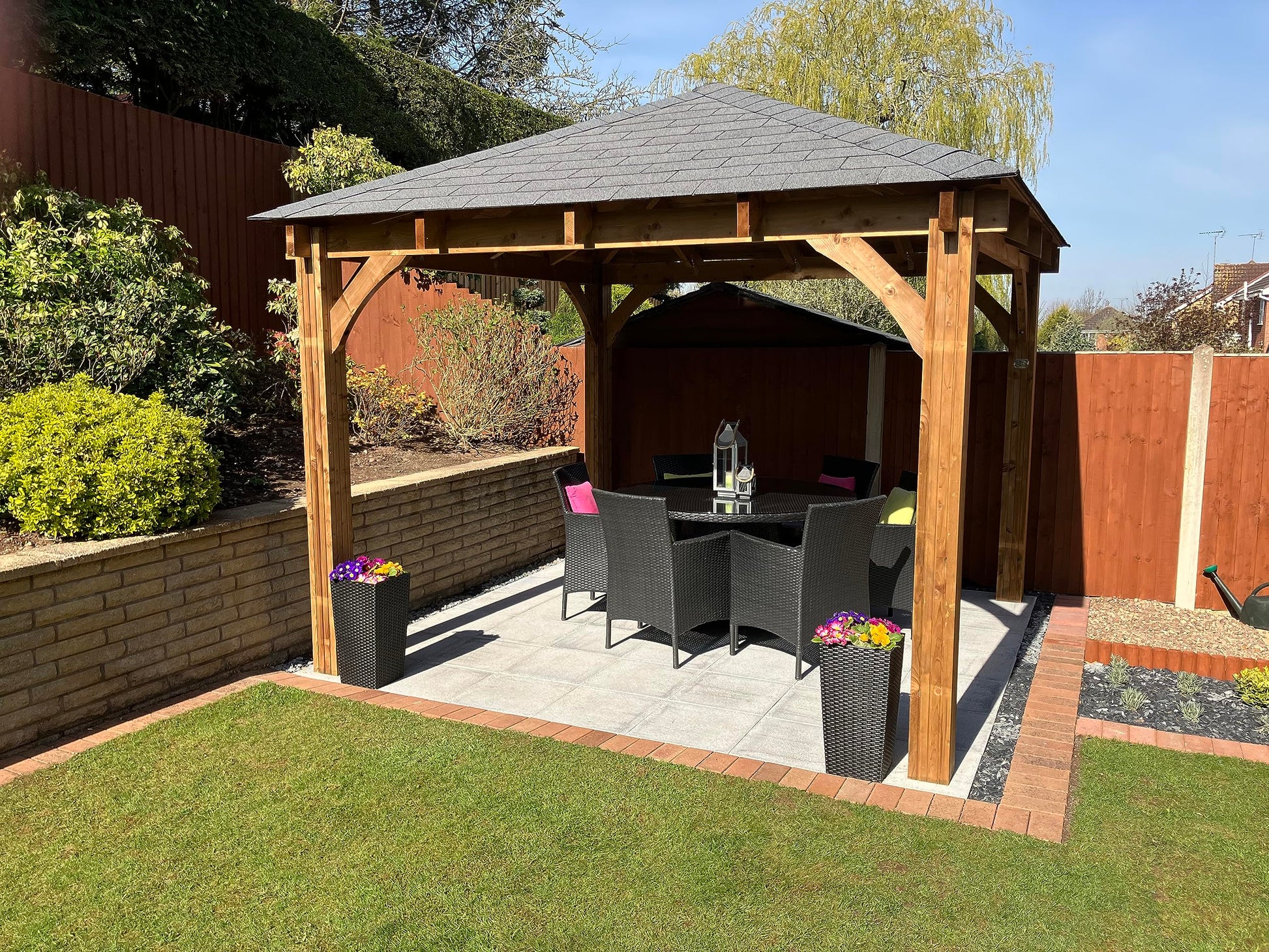 Garden installation of the gazebo over a patio dining set with planters, low brick wall and wooden fence on a sunny day